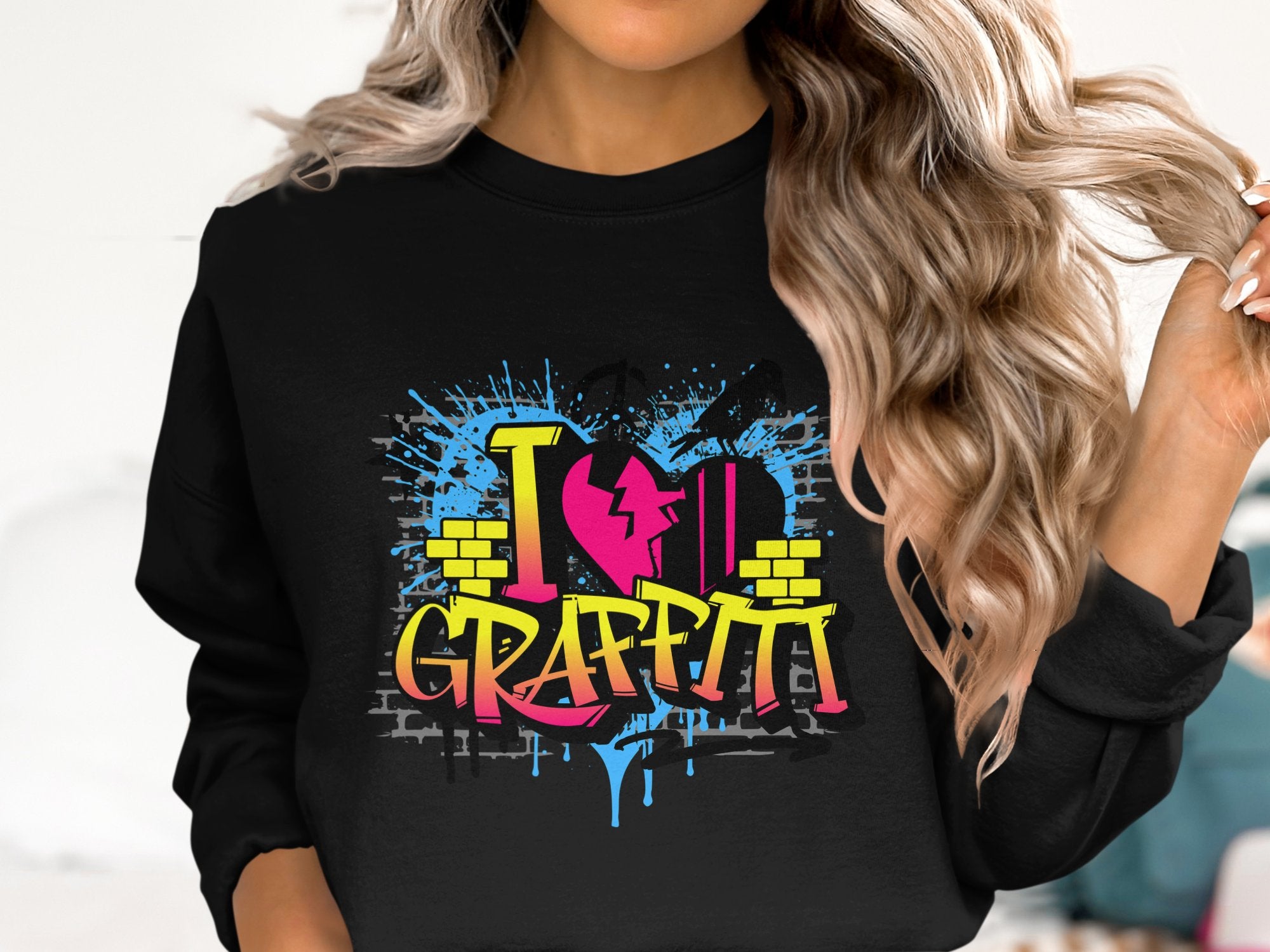 Someone with long, wavy hair wears a Garment Graphics sweatshirt adorned with a vibrant graffiti design featuring a heart, lightning bolt, and I Love Graffiti in blue, pink, and yellow splashes, capturing an urban aesthetic.