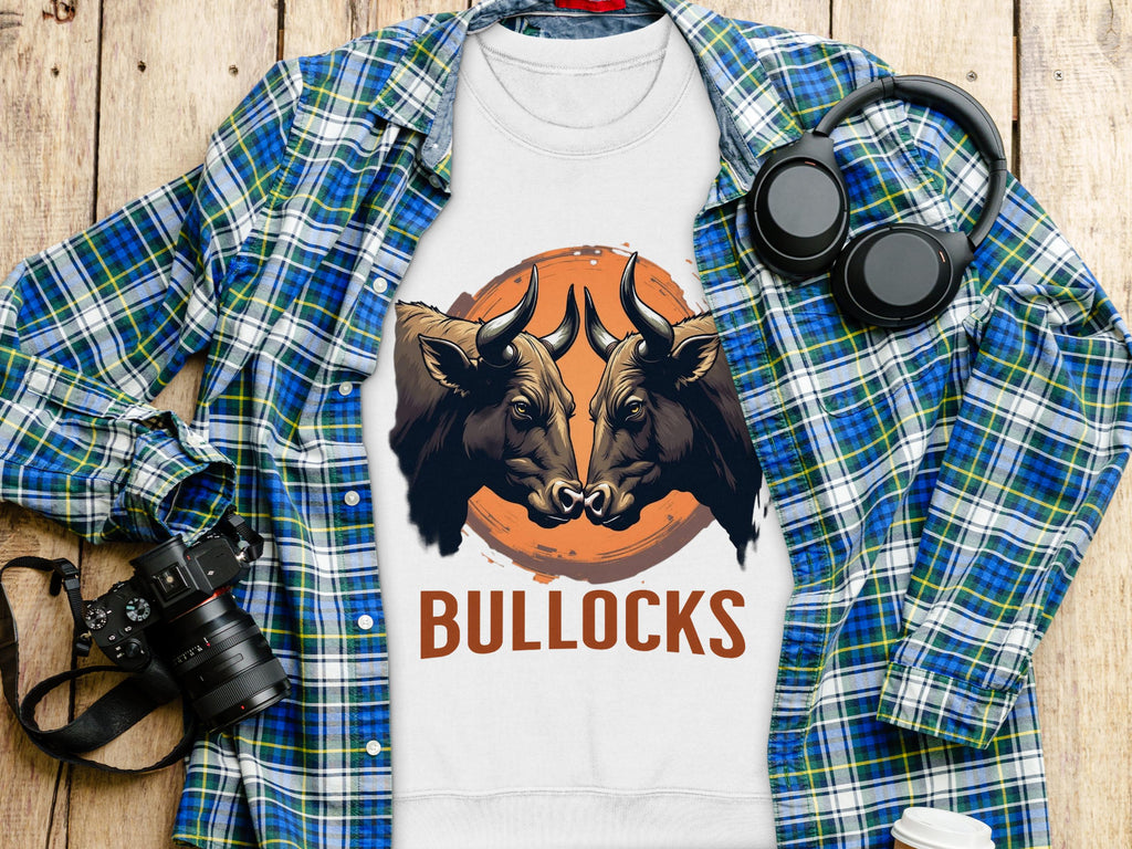 Flat lay of a Garment Graphics white t-shirt with a bold BULLOCKS design, featuring two bulls. The unisex shirt is displayed on wood next to a blue plaid shirt, headphones, camera, and white cup.