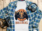 Flat lay of a Garment Graphics white t-shirt with a bold BULLOCKS design, featuring two bulls. The unisex shirt is displayed on wood next to a blue plaid shirt, headphones, camera, and white cup.