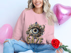 A person in a Garment Graphics pink sweatshirt with an eye design and GAZE text holds a red rose, surrounded by pink heart balloons. They wear ripped jeans, and their long, wavy blonde hair adds charm to this unisex look.