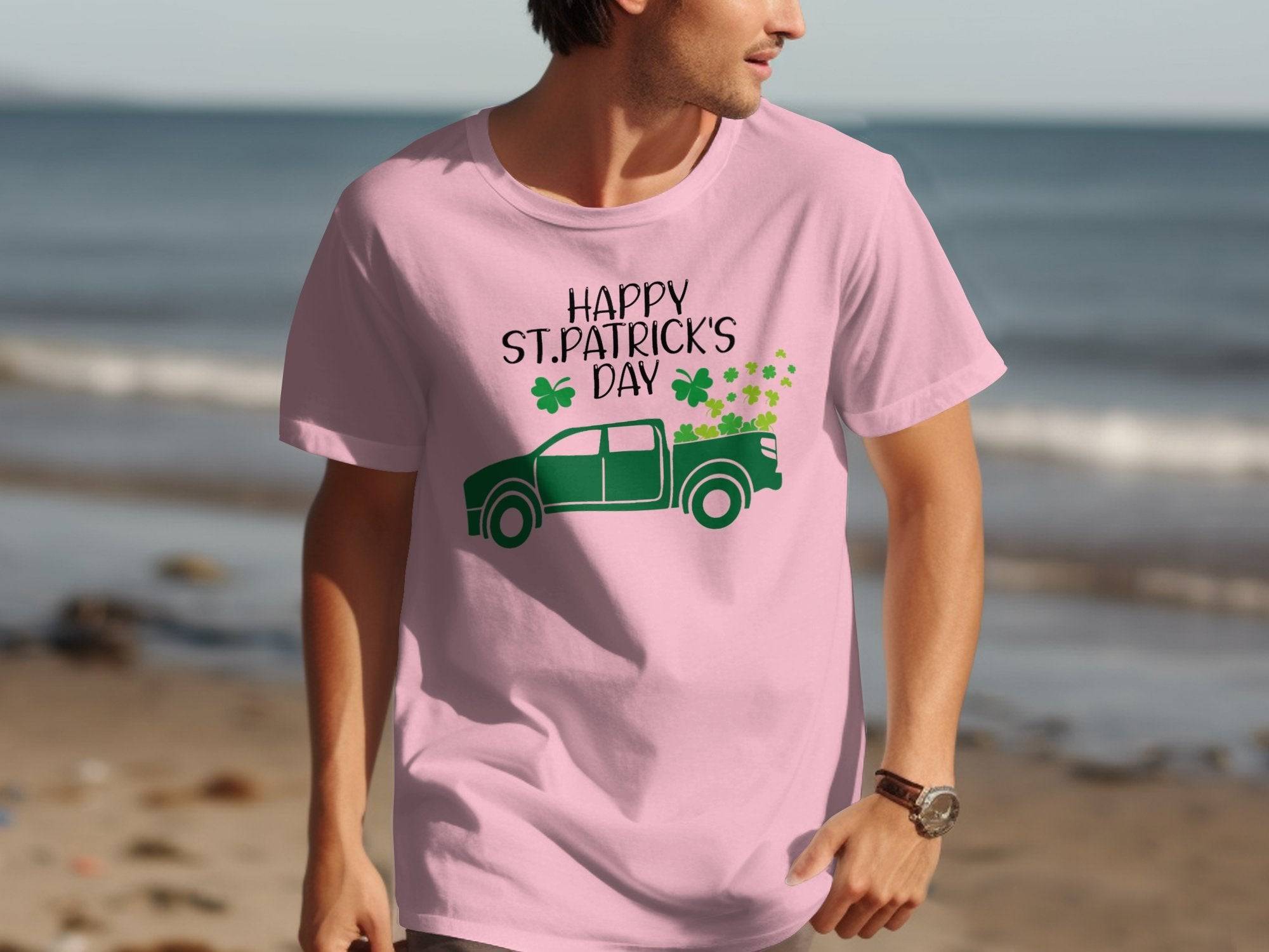 Wearing a Garment Graphics pink T-shirt adorned with a green pickup truck and clovers design, along with the Happy St. Patricks Day text, someone walks on the beach as waves gently touch the sand in the backdrop, showcasing their Irish pride.