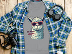 A Garment Graphics unisex gray sweatshirt with a llama in red sunglasses and No Prob-llama text is placed on a plaid shirt. Nearby are a camera, headphones, and takeaway coffee on wood. Its perfect for adding humor to any outfit.