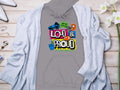 A Garment Graphics gray hoodie with a vibrant Hey Ho Lets Go! Loud & Proud design in mixed fonts pairs stylishly with a light gray cardigan, displayed on wood alongside white flowers, a ring, and perfume—ideal for making bold statements.