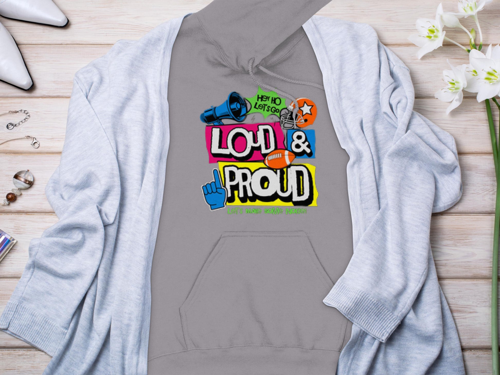 A Garment Graphics gray hoodie with a vibrant Hey Ho Lets Go! Loud & Proud design in mixed fonts pairs stylishly with a light gray cardigan, displayed on wood alongside white flowers, a ring, and perfume—ideal for making bold statements.