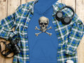 A blue Garment Graphics streetwear hoodie with a skull and crossbones design lies on wood, partly covered by a blue-green plaid shirt. Nearby are headphones, a camera, a takeaway coffee cup, and a phone in view.