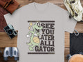 Garment Graphics presents a gray casual t-shirt featuring a cartoon alligator with a basketball and star near its eye. The playful See You Later Alligator text adds charm. Its styled on wood, surrounded by a camera, binoculars, and leather accessories.