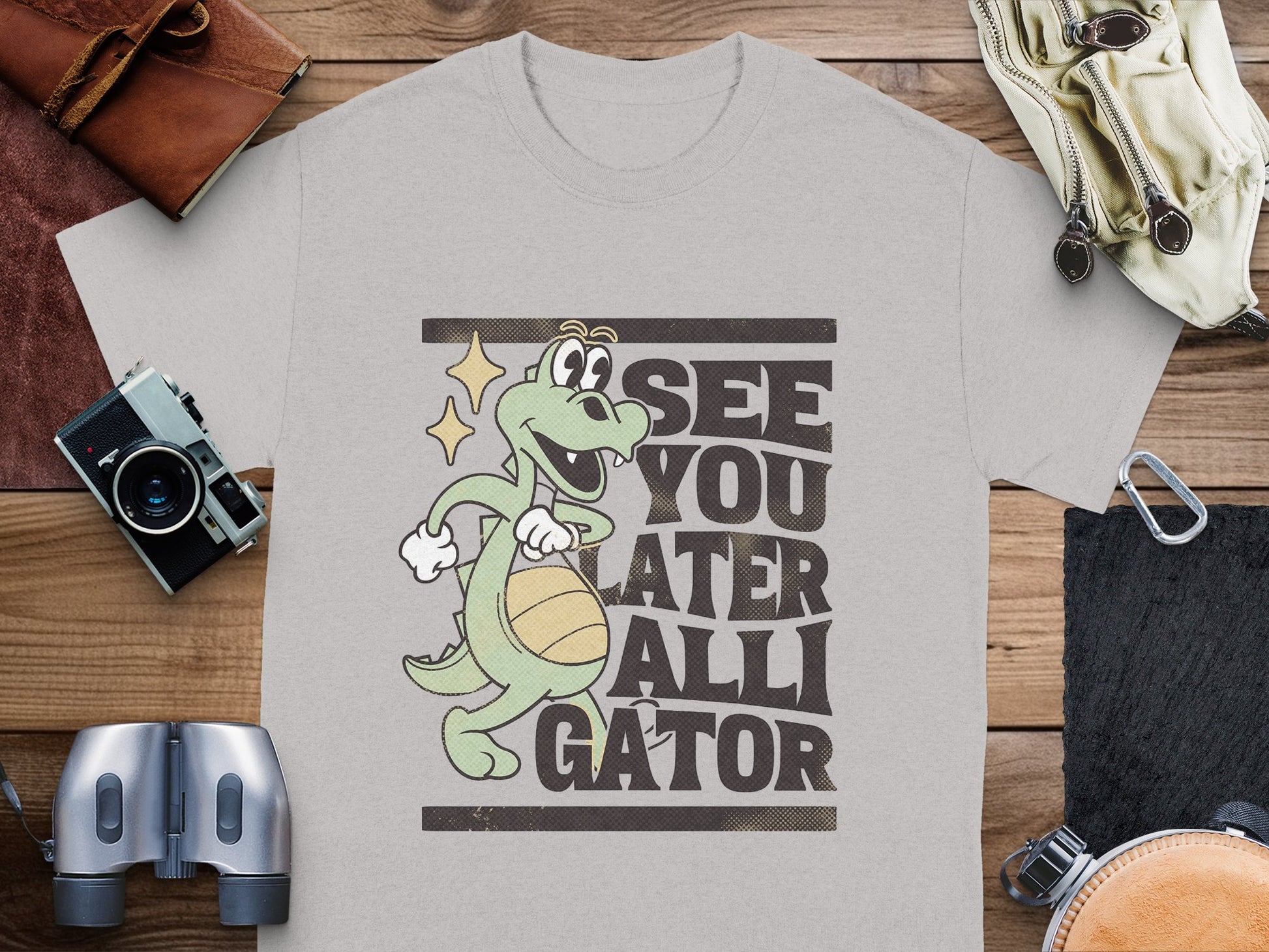 Garment Graphics presents a gray casual t-shirt featuring a cartoon alligator with a basketball and star near its eye. The playful See You Later Alligator text adds charm. Its styled on wood, surrounded by a camera, binoculars, and leather accessories.