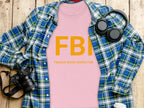 Garment Graphics pink FBI Female Boob Inspector gag gift T-shirt in womens sizing is placed over a blue and green plaid shirt, with headphones, a camera, a white cup, and a black smartphone nearby on the wooden surface.
