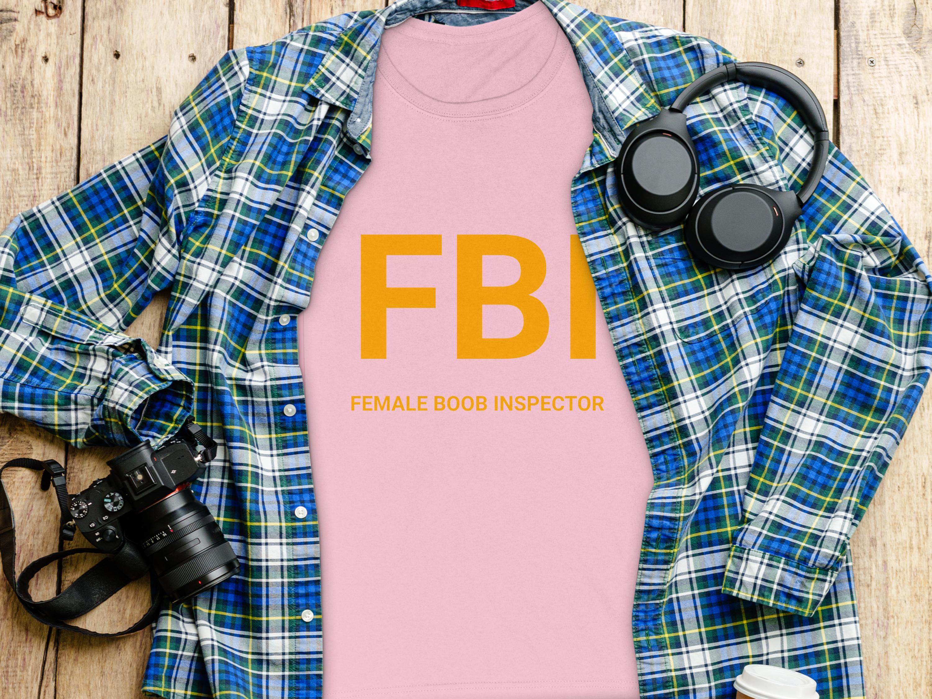Garment Graphics pink FBI Female Boob Inspector gag gift T-shirt in womens sizing is placed over a blue and green plaid shirt, with headphones, a camera, a white cup, and a black smartphone nearby on the wooden surface.