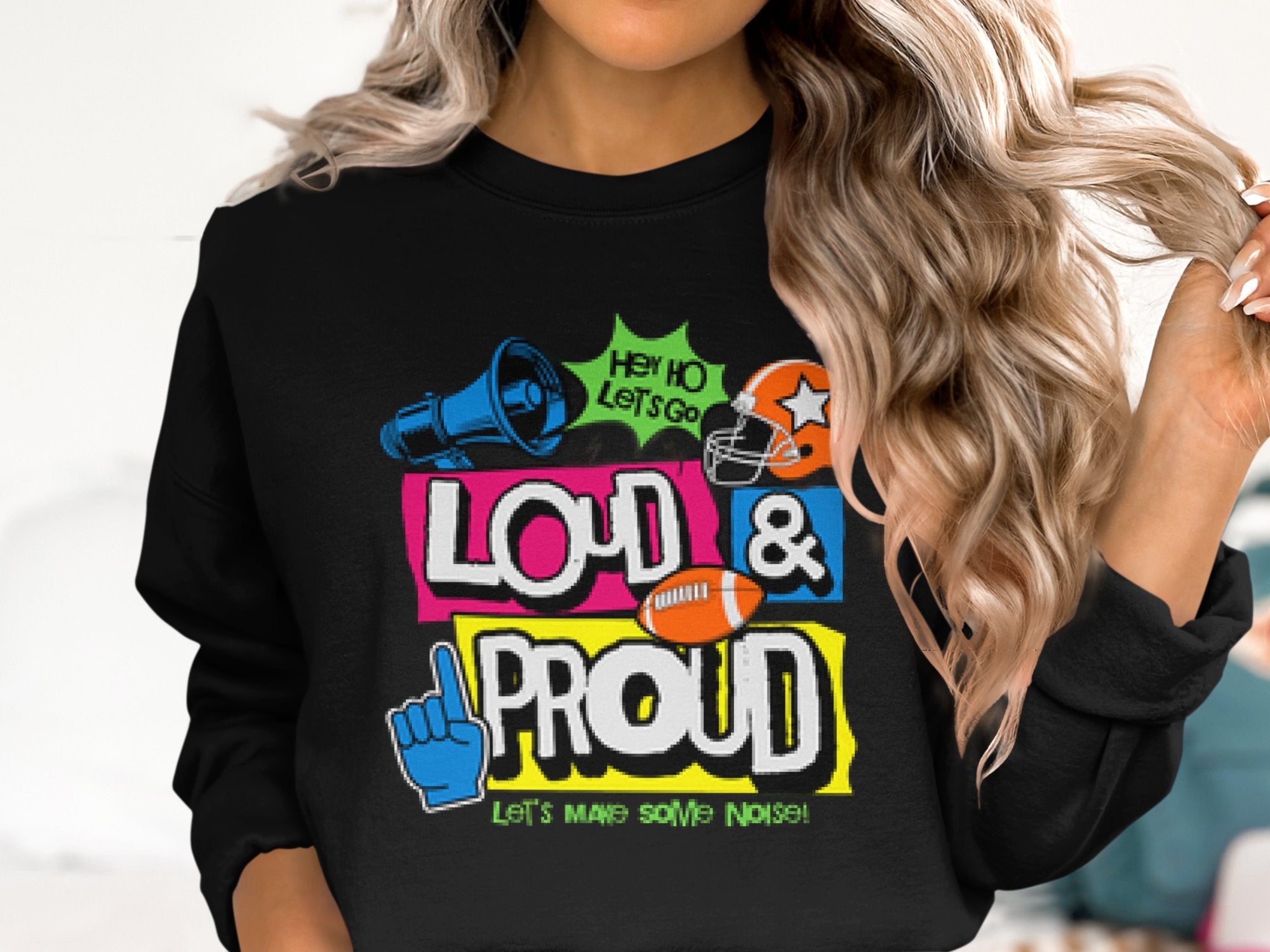 A person with long, wavy hair is wearing a Garment Graphics Loud and Proud sweatshirt. The black design features vibrant graphics and text reading LOUD & PROUD, along with a megaphone, foam finger, football, and helmet images. Ideal for those looking for unisex sizing.