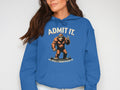 A person wears a Garment Graphics classic fit blue hoodie featuring a Bigfoot character with two fingers raised, alongside the text Admit It. Life would be boring without me. The individual, with long dark hair, is only partially visible.