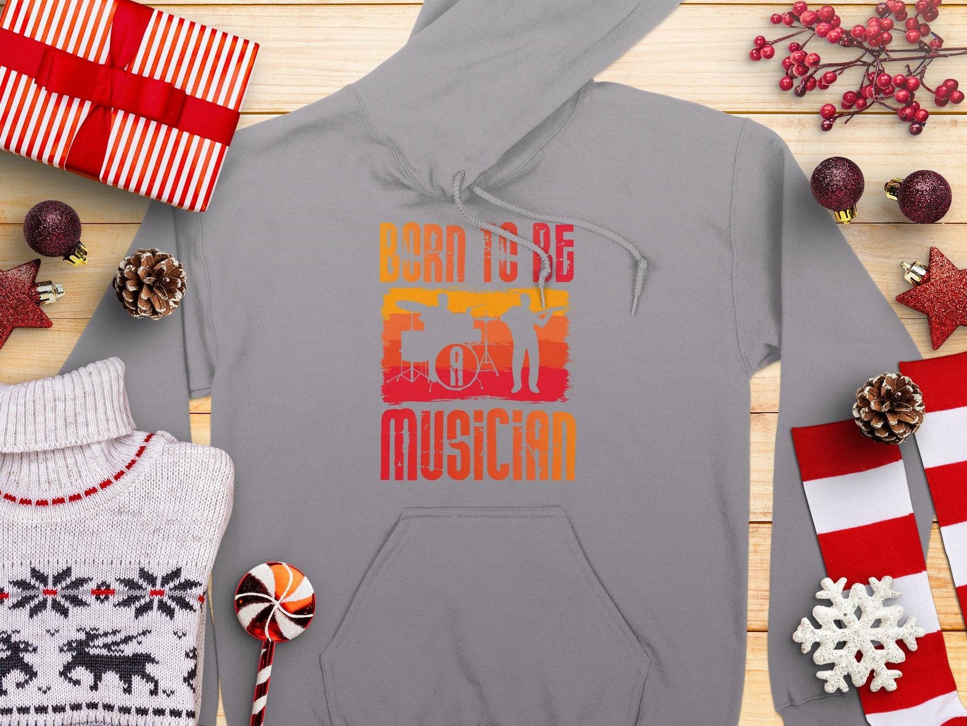 The Garment Graphics unisex hoodie with Born to be a Musician and a drummers silhouette is ideal for music lovers, surrounded by festive items like wrapped gifts, pinecones, candy canes, and a cozy reindeer-patterned sweater on a wooden surface.