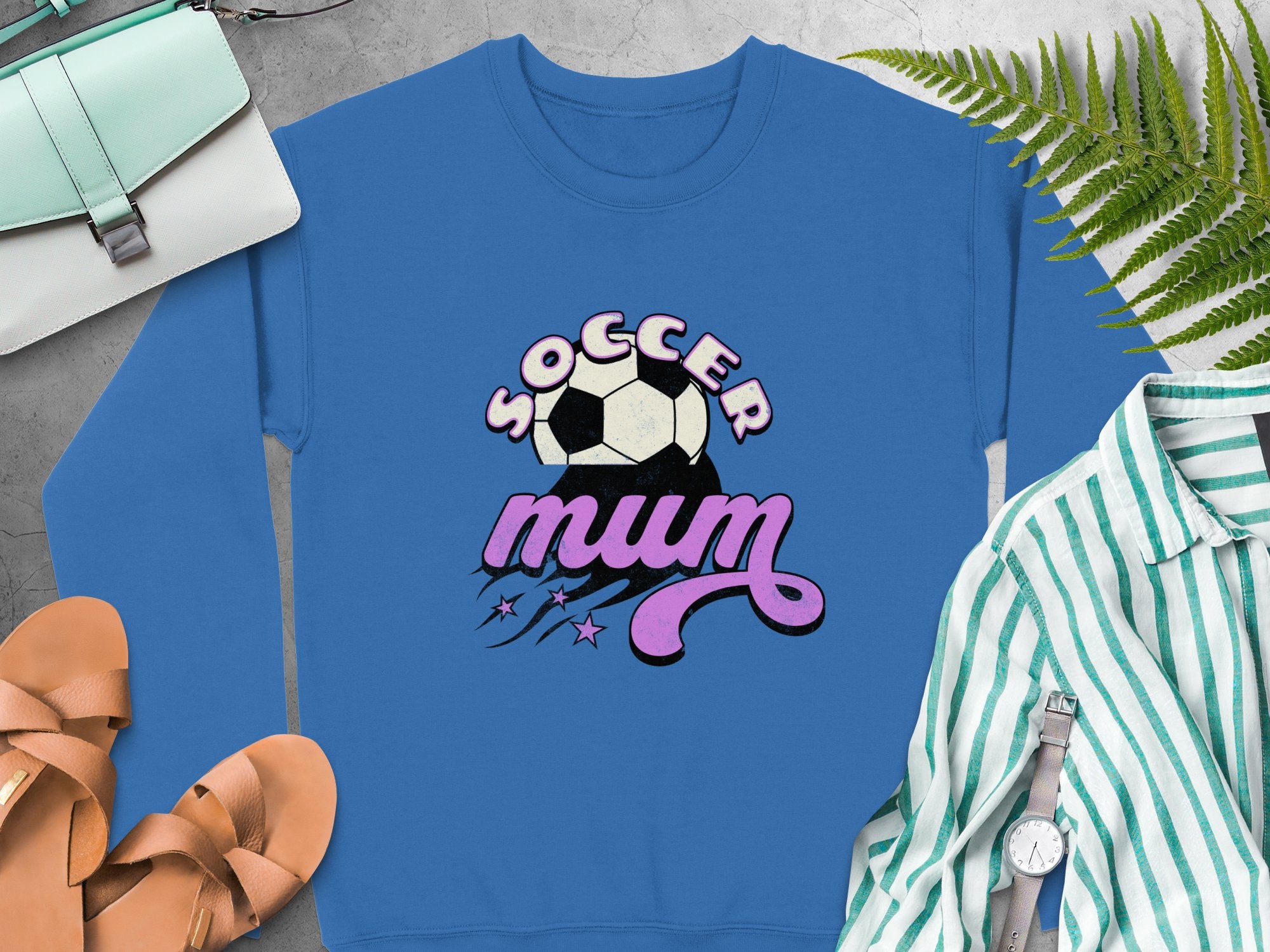 A Garment Graphics blue sweatshirt with a vintage Soccer Mum soccer ball design is displayed on a concrete surface alongside a striped shirt, purse, sandals, watch, and fern.