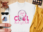 A Garment Graphics white sweatshirt with Just be Cool Rock and Roll text and pink headphones lies on wood. Fashion lovers will like nearby items: a yellow jacket, makeup brushes, eyeshadow palette, sunglasses, pink shoes, nail polish, and a blue pouch.