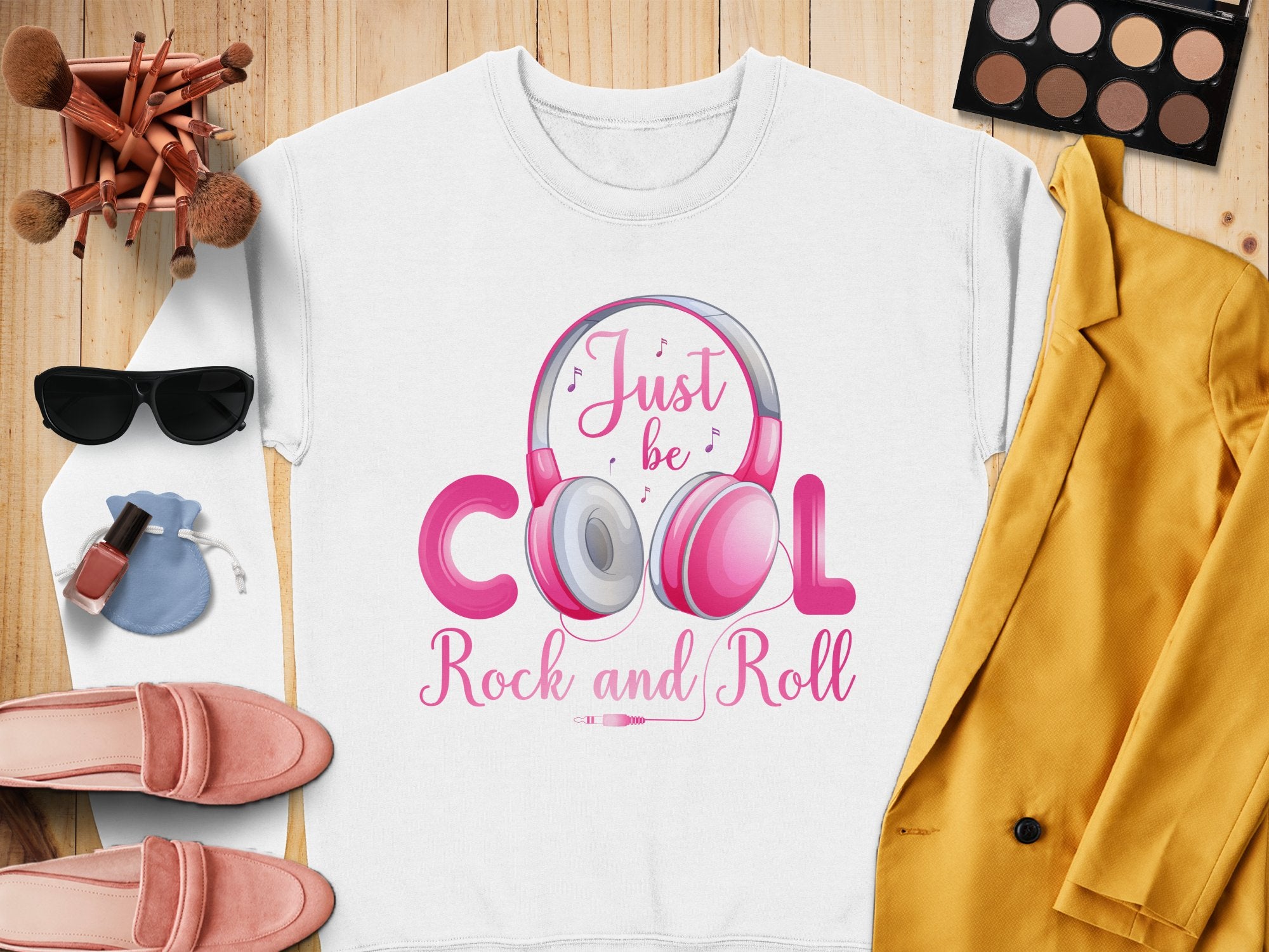 A Garment Graphics white sweatshirt with Just be Cool Rock and Roll text and pink headphones lies on wood. Fashion lovers will like nearby items: a yellow jacket, makeup brushes, eyeshadow palette, sunglasses, pink shoes, nail polish, and a blue pouch.