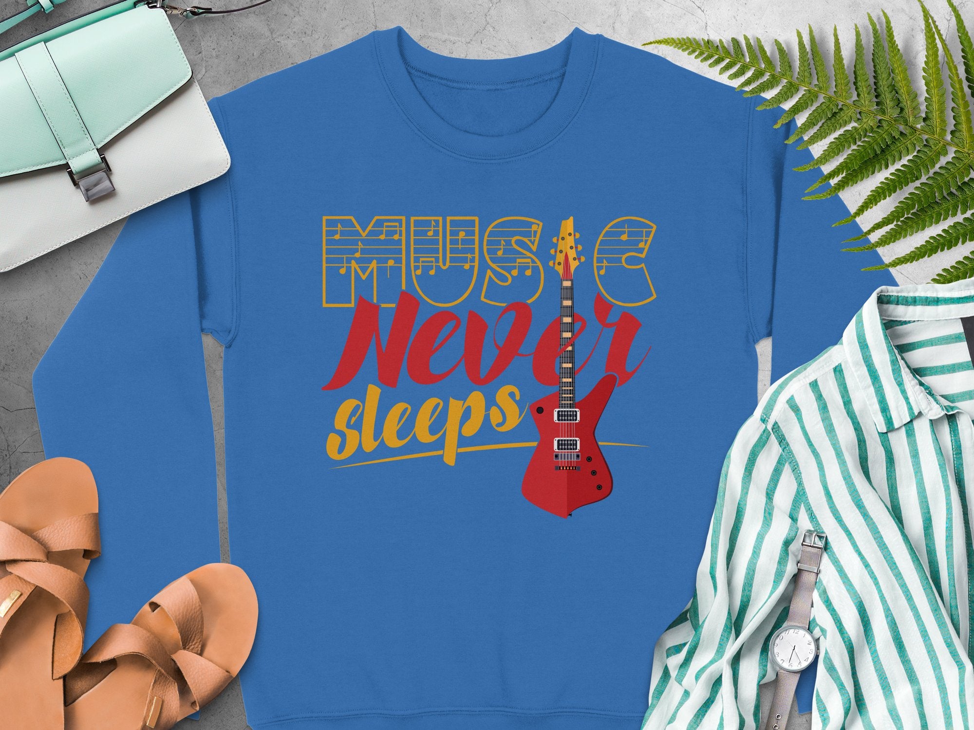 Discover Garment Graphics trendy blue sweatshirt, showcasing a vibrant Music Never Sleeps Guitar design with a red electric guitar. Ideal for music lovers, its styled with a green-striped shirt, sandals, and teal purse on a gray backdrop featuring a fern and wristwatch.