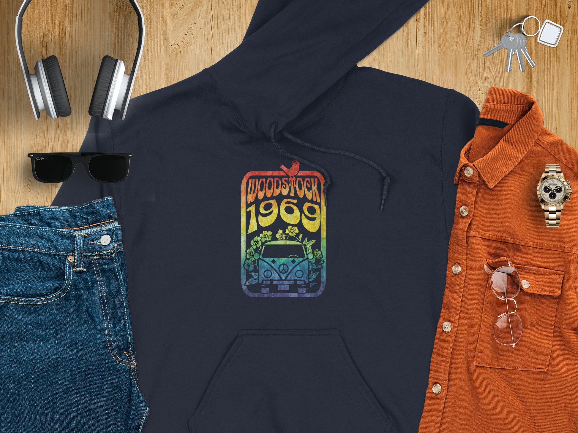 A retro black Garment Graphics hoodie featuring a vibrant Woodstock 1969 design lies on wood, surrounded by jeans, headphones, sunglasses, a watch, keys, an orange button-up shirt, and glasses.