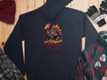 A Garment Graphics black hoodie with a Turkey On The Run cartoon design sits on a wooden surface, accompanied by a maroon knit hat, gray and black gloves, a plaid scarf, and a green and red plaid shirt for the perfect cozy look.