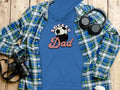 A flat lay image features a Garment Graphics blue hoodie with Soccer Dad print, alongside a plaid shirt, headphones, camera, and coffee cup lid on a wooden surface.