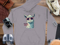 The Garment Graphics classic fit gray hoodie showcases an illustrated llama in sunglasses with No Prob-Llama, surrounded by travel items like a camera, binoculars, a bag, and books on a wooden surface.