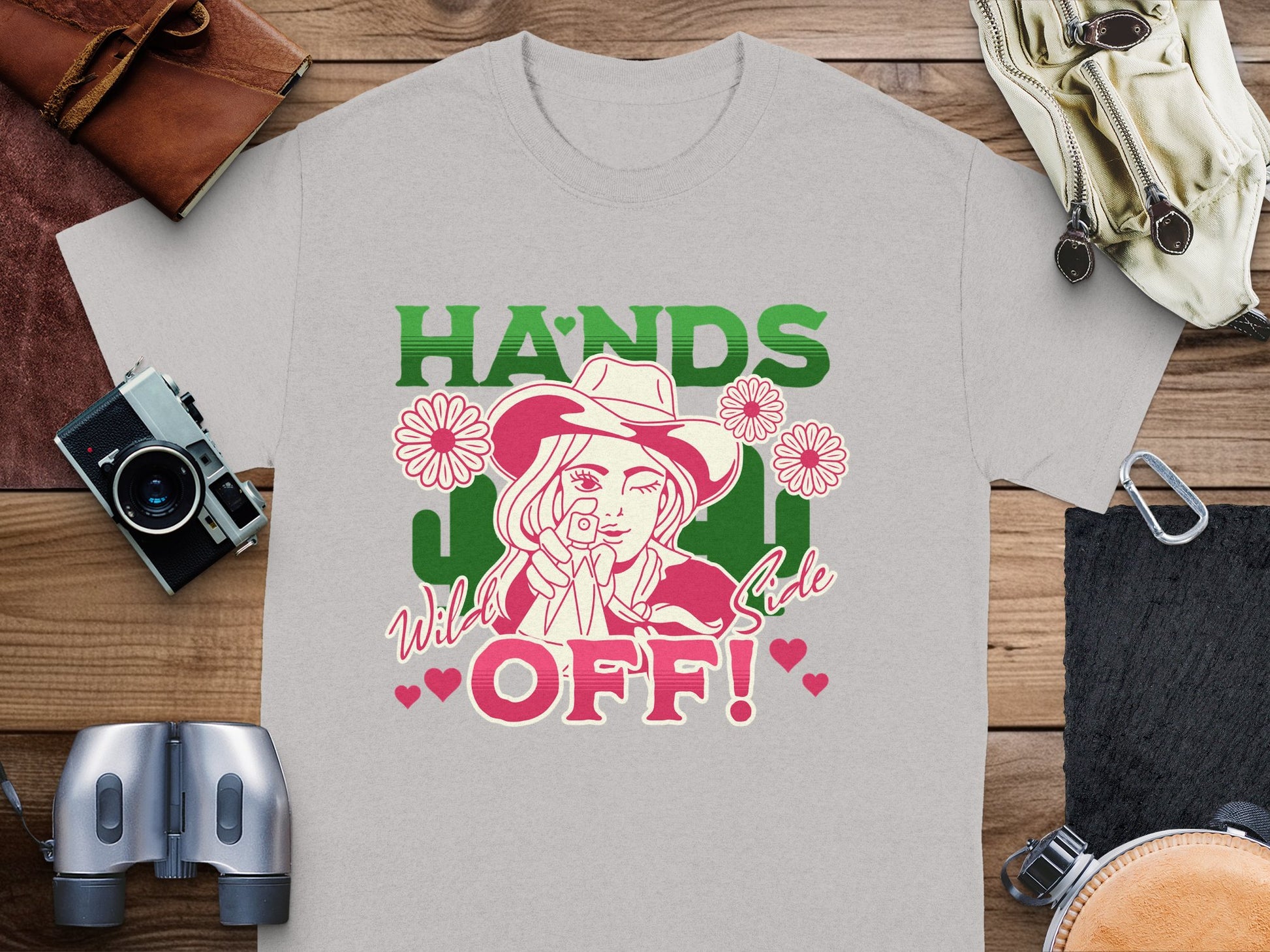 The Garment Graphics gray T-shirt showcases a bold cowgirl with a hat and gun, surrounded by flowers. It reads Hands Off! Wild Side. The classic fit shirt is displayed on wood with cameras, a book, binoculars, and gloves. Ideal for Hands Off T-Shirts enthusiasts.