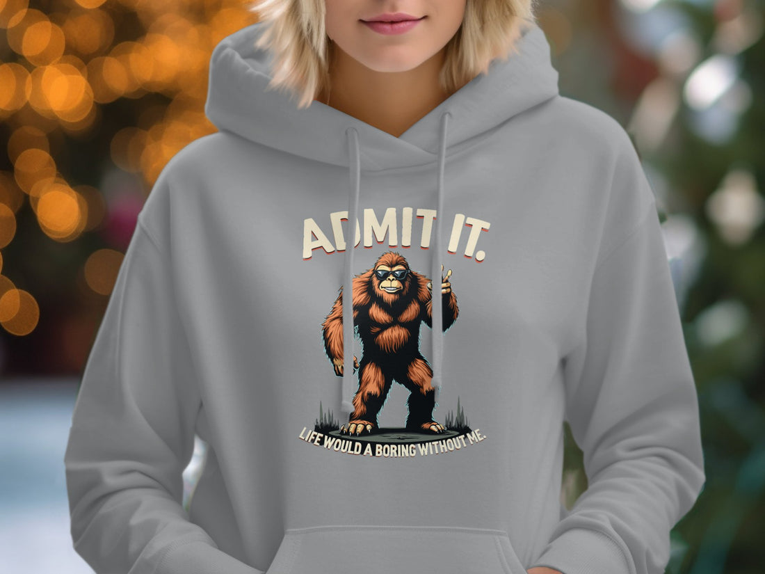 Garment Graphics white classic-fit hoodie features a Bigfoot illustration with Admit It, Life Would Be Boring Without Me. Its displayed on a wooden surface with a straw hat, sunglasses, sandals, notebook, map, and camera—a cool character ensemble.