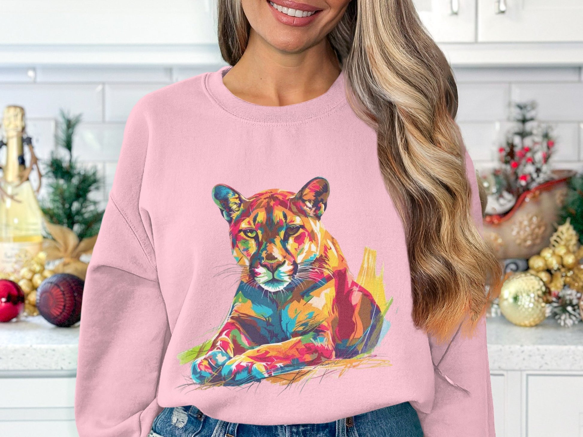 A person wearing a Garment Graphics pink sweatshirt with a vibrant cougar design. The unique style complements the festive decorations and white tiled wall, highlighting its unisex sizing for everyone to enjoy.