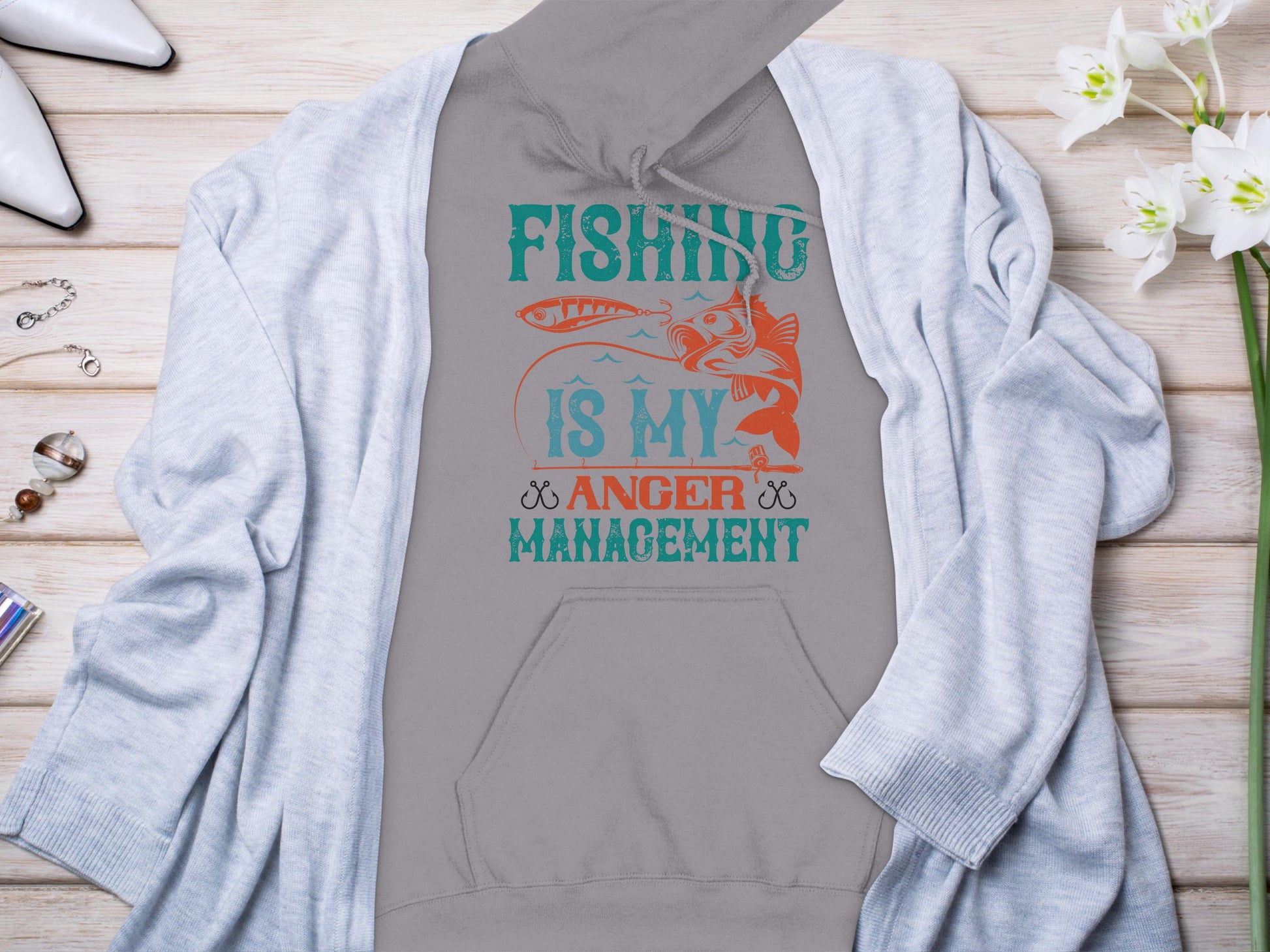 Garment Graphics presents a gray hoodie reading Fishing is my anger management with a teal and orange fish graphic. Styled for the anglers wardrobe, its set on wood alongside a light gray cardigan, white heels, flowers, and jewelry.