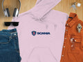 A pink hoodie with the iconic Scania logo, designed by Garment Graphics, is centered on a wooden surface. Its surrounded by headphones, sunglasses, blue jeans, a rust-colored shirt, a silver watch, and keys with a small white house-shaped keychain.