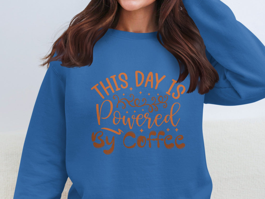 A person wears a Garment Graphics sweatshirt with the vibrant orange script, This day is powered by coffee. Their brown, wavy hair falls to their shoulders against a light, neutral background.