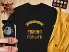 A Garment Graphics black graphic sweatshirt with the yellow text Father and Son Fishing for Life sits on a wooden surface, alongside a mustard blazer, makeup brushes, an eyeshadow palette, sunglasses, pink shoes, and a small blue pouch.