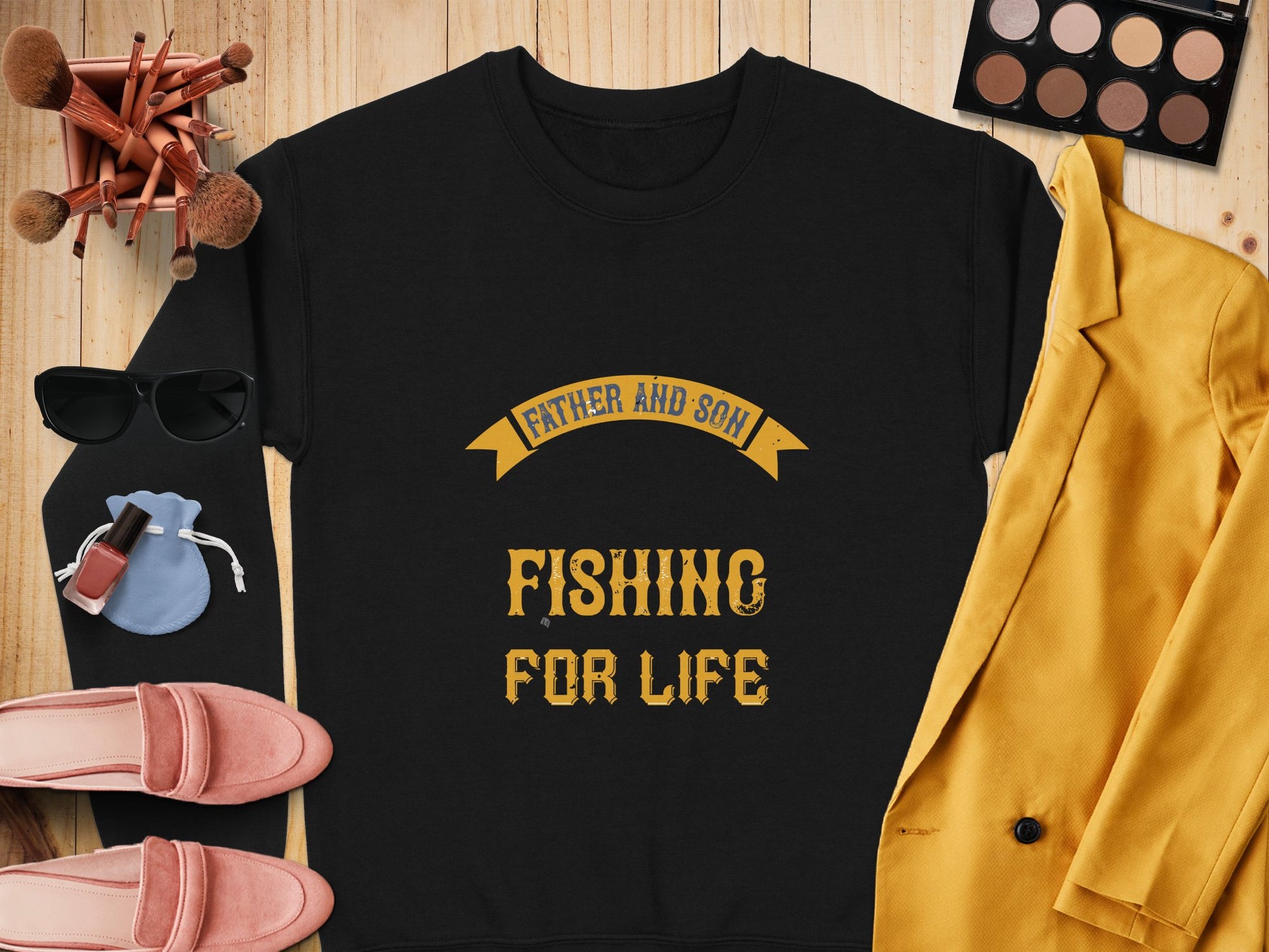 A Garment Graphics black graphic sweatshirt with the yellow text Father and Son Fishing for Life sits on a wooden surface, alongside a mustard blazer, makeup brushes, an eyeshadow palette, sunglasses, pink shoes, and a small blue pouch.