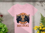 On a concrete backdrop, this Garment Graphics pink T-shirt showcases bold graphics of two bulls with an orange circle, and BULLOCKS below. Green plants and yellow flowers add a natural element to the striking design.