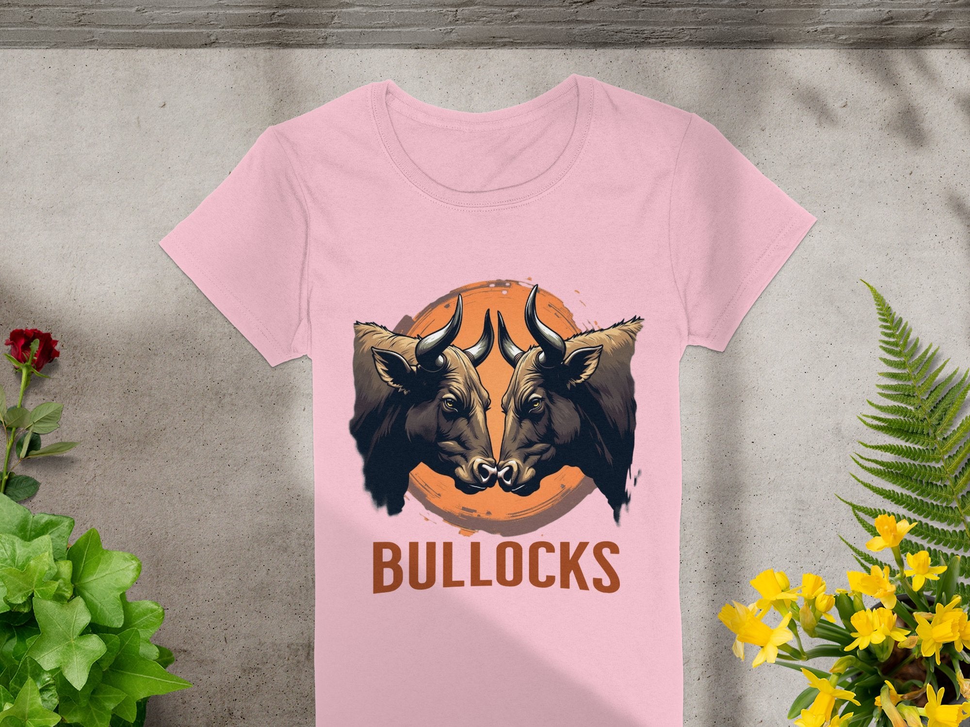 On a concrete backdrop, this Garment Graphics pink T-shirt showcases bold graphics of two bulls with an orange circle, and BULLOCKS below. Green plants and yellow flowers add a natural element to the striking design.