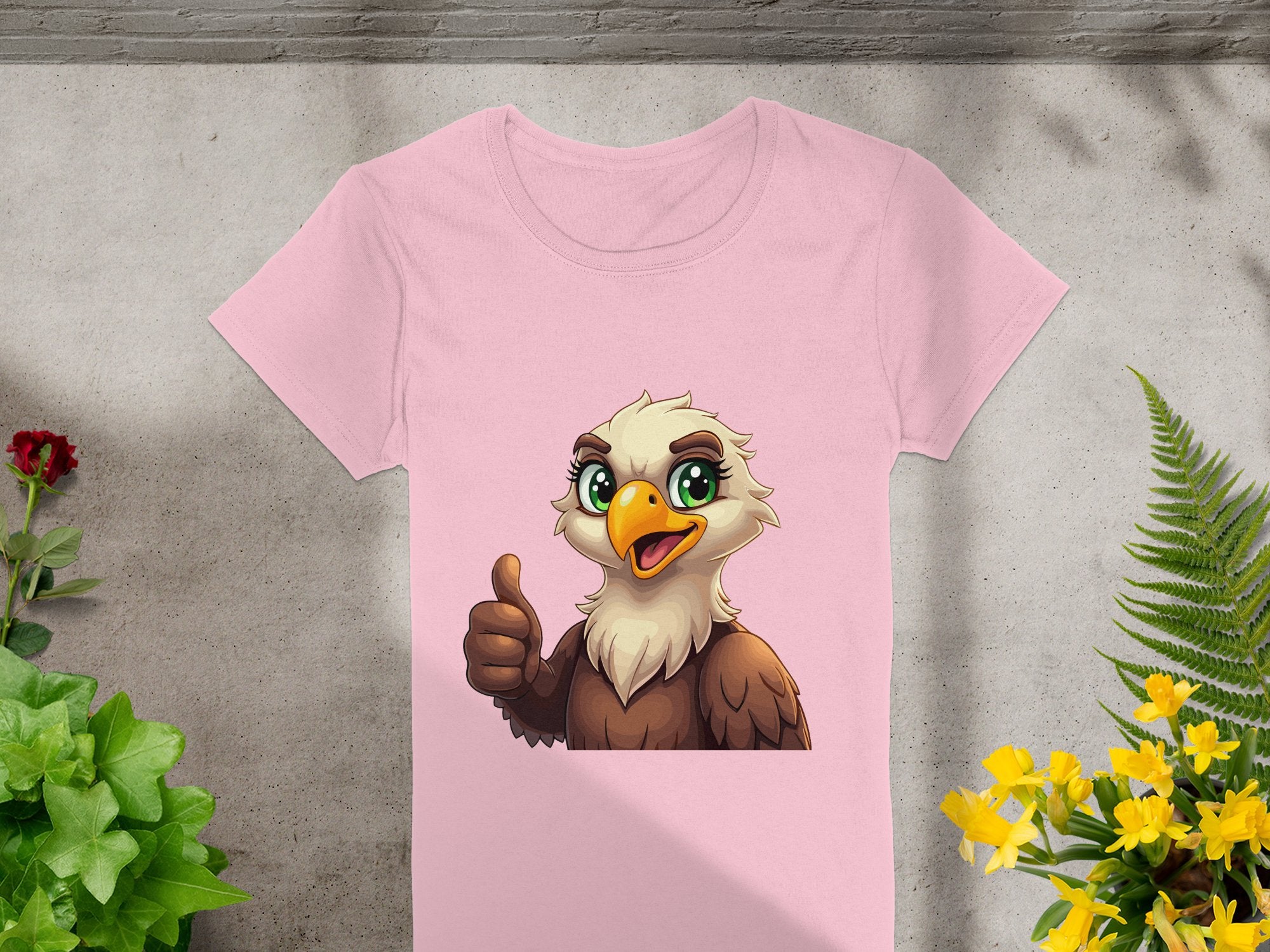 Garment Graphics presents a pink cartoon T-shirt featuring a cheerful eagle with big green eyes giving a thumbs-up. Ideal for animal lovers, this vibrant shirt is displayed flat on concrete, surrounded by colorful red, green, and yellow plants.