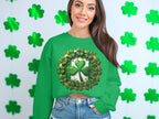 A person with long hair smiles while wearing a Garment Graphics green sweatshirt featuring a lucky four-leaf clover. They stand in front of a wall adorned with green shamrocks, capturing the Irish spirit and St. Patricks Day theme.