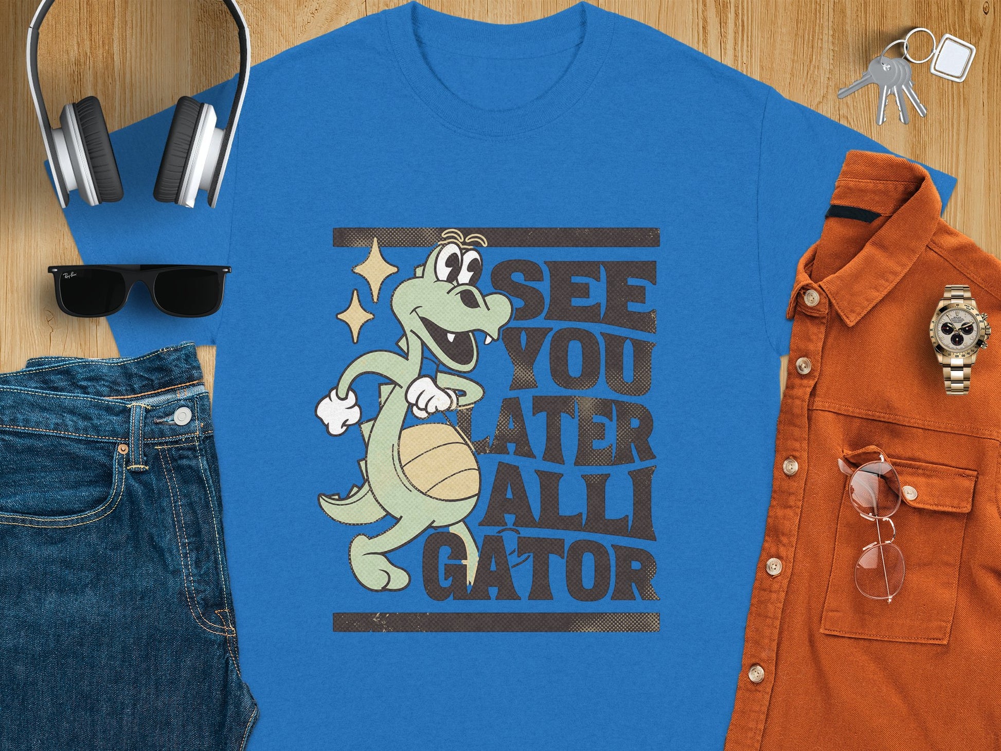 A flat lay image displays a blue T-shirt by Garment Graphics, featuring a cartoon alligator with a basketball and the phrase See You Later Alligator. Ideal for casual wear, it’s surrounded by headphones, keys, sunglasses, jeans, an orange shirt, and a watch.