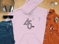 Garment Graphics presents a pink retro 45 RPM music hoodie on a wooden surface. Ideal for vinyl enthusiasts, its accompanied by headphones, sunglasses, keys, jeans, an orange button-up shirt, and a wristwatch.