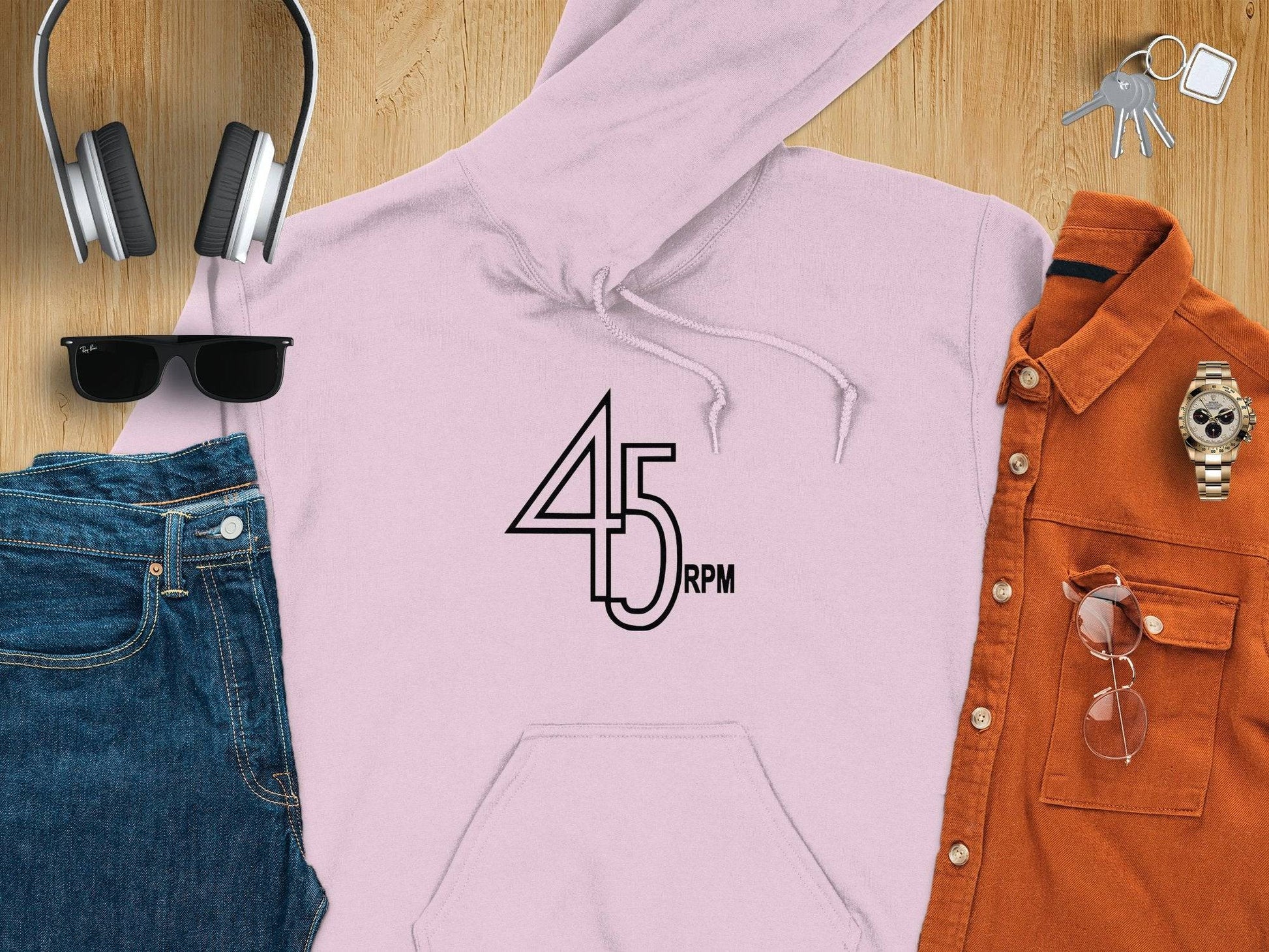 Garment Graphics presents a pink retro 45 RPM music hoodie on a wooden surface. Ideal for vinyl enthusiasts, its accompanied by headphones, sunglasses, keys, jeans, an orange button-up shirt, and a wristwatch.