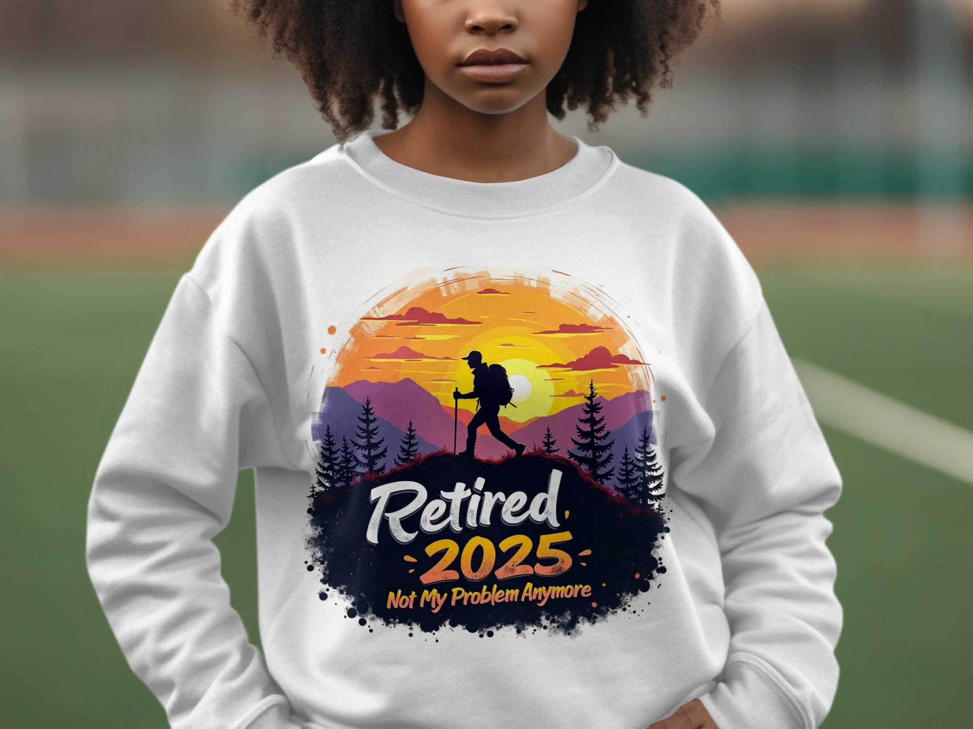 A person in a white Garment Graphics retirement sweatshirt, unisex-sized, shows a hiker against a sunset with the text Retired 2025, Not My Problem Anymore, set against a blurred outdoor sports field backdrop.