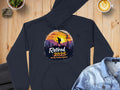 The black hiking hoodie by Garment Graphics, showcases a trekker against a sunset mountain design with the text Retired 2025: Not My Problem Anymore, styled on wood next to denim, a suit piece, coffee, and plant.