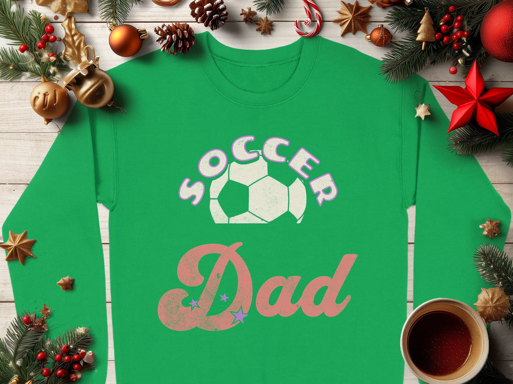 A green Garment Graphics Soccer Dad sweatshirt featuring a soccer ball graphic is on a wooden surface amid festive holiday decorations like pine branches, ornaments, and pine cones. A steaming cup of hot beverage complements the cozy scene. The sweatshirt offers unisex sizing for an ideal fit.