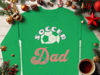A green Garment Graphics Soccer Dad sweatshirt featuring a soccer ball graphic is on a wooden surface amid festive holiday decorations like pine branches, ornaments, and pine cones. A steaming cup of hot beverage complements the cozy scene. The sweatshirt offers unisex sizing for an ideal fit.