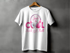 A white Garment Graphics T-shirt on a hanger displays a pink and silver graphic of playful headphones with Just be Cool Rock and Roll in stylized text, ideal for music lovers. Its set against a textured gray wall.