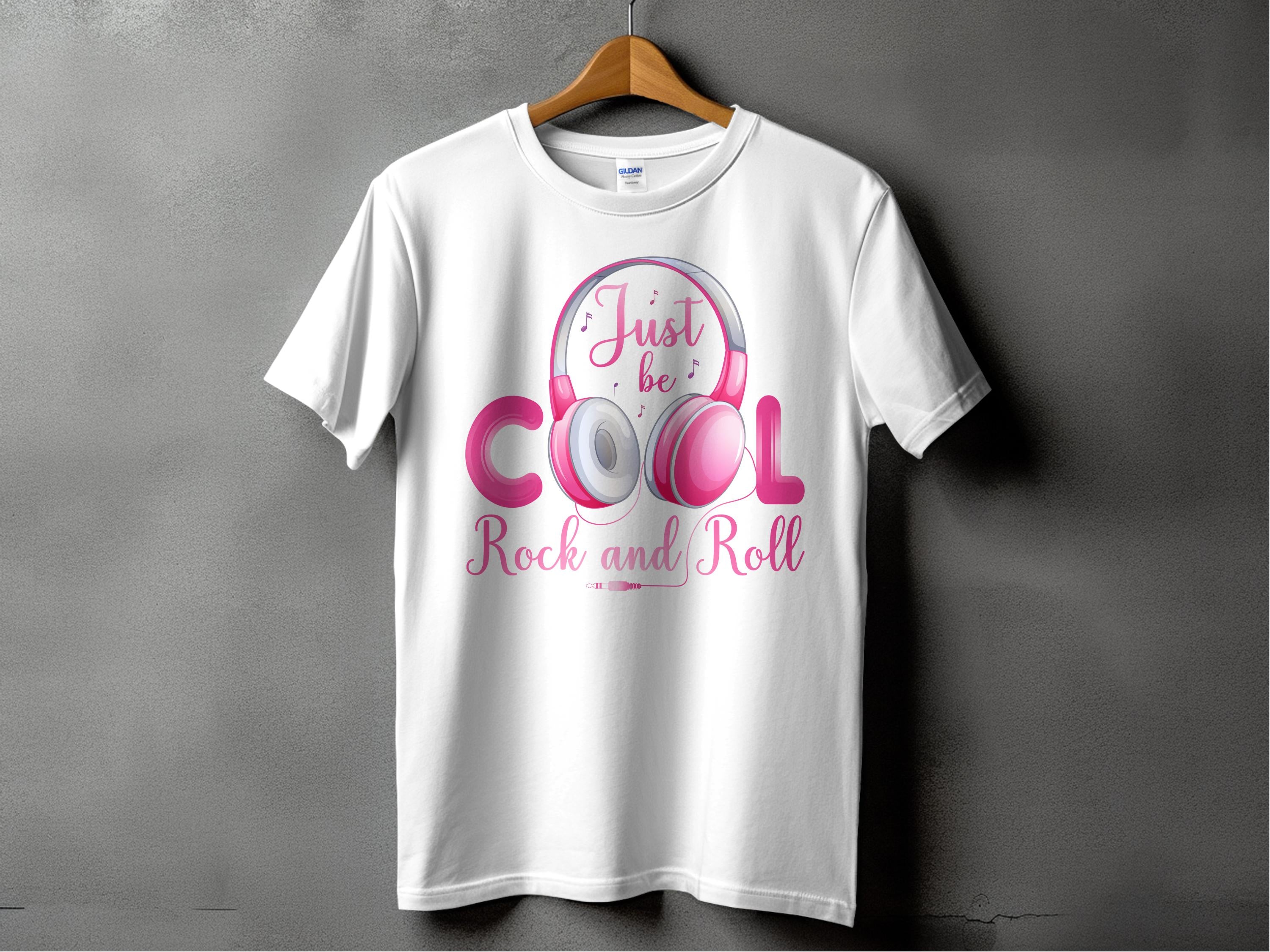 A white Garment Graphics T-shirt on a hanger displays a pink and silver graphic of playful headphones with Just be Cool Rock and Roll in stylized text, ideal for music lovers. Its set against a textured gray wall.