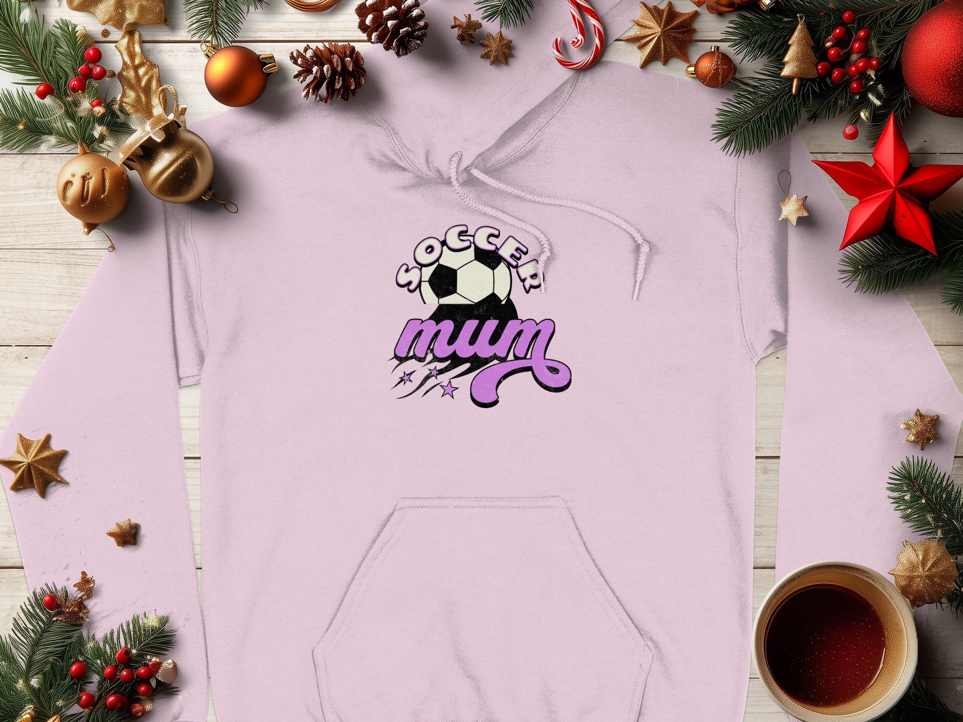 This cozy Soccer Mum hoodie by Garment Graphics, in light pink with a classic fit and purple accents, is beautifully showcased among Christmas decor, featuring pine branches, ornaments, pinecones, and a steaming coffee cup on a wooden surface.