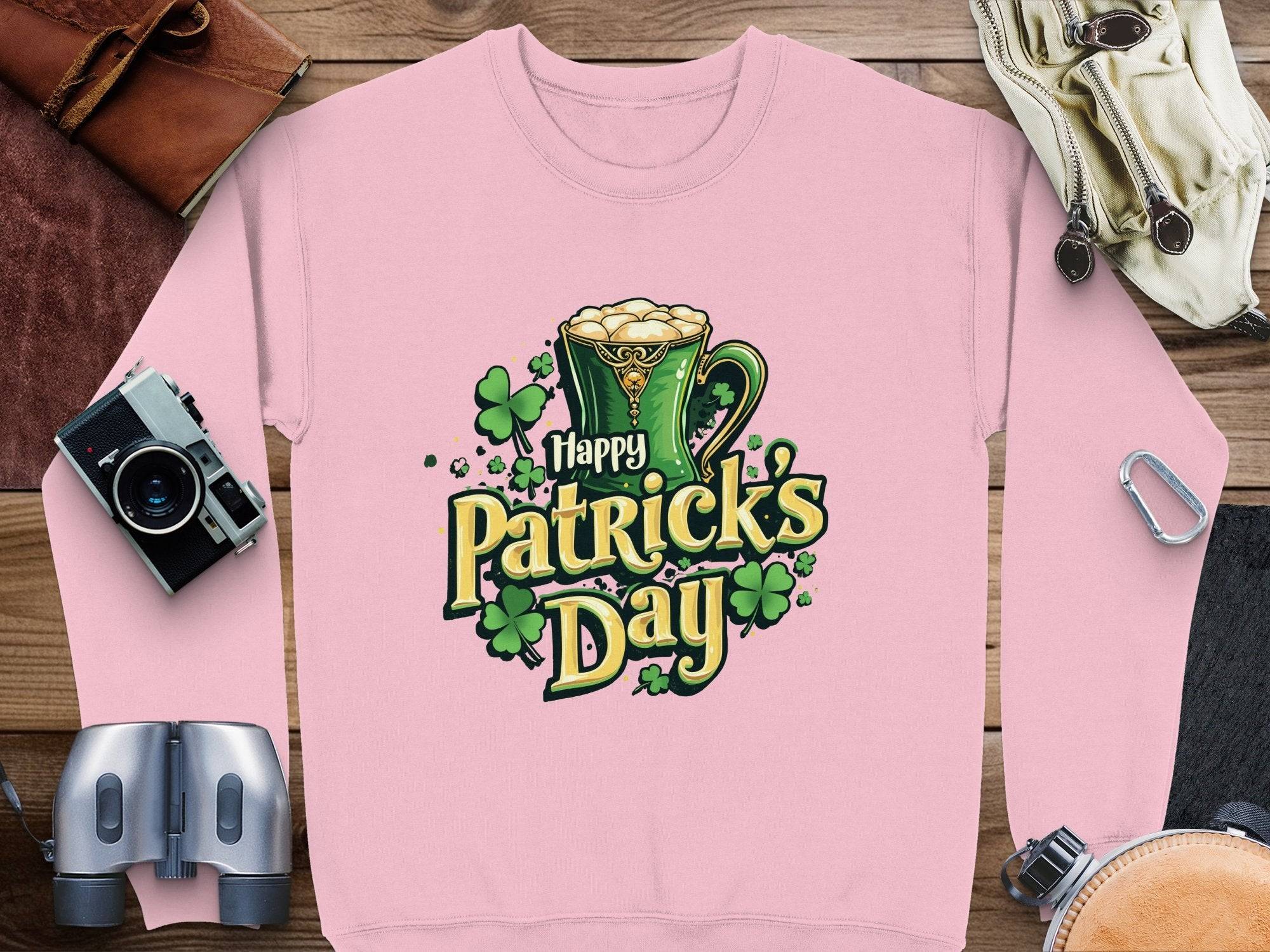 Garment Graphics offers a pink sweatshirt perfect for St. Patricks Day. It showcases a festive green beer mug, shamrocks, and Happy Patricks Day text on a vivid background. Laid out on wood, this piece captures the holiday spirit effortlessly.