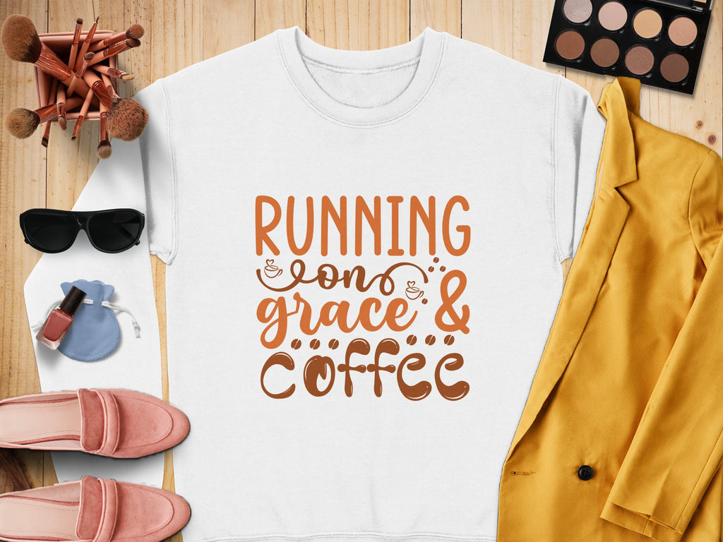 A white Garment Graphics sweatshirt with Running on Grace & Coffee in bold orange is styled with a yellow blazer, pink shoes, sunglasses, makeup brushes, and a beige eyeshadow palette. It offers unisex sizing for versatile wear on the wooden surface.