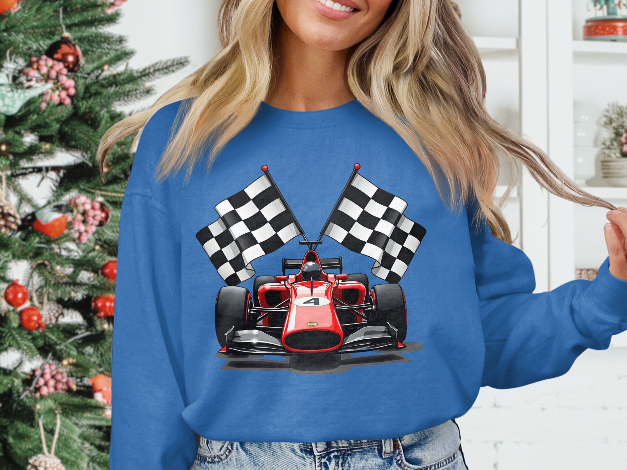 A woman wears a vibrant blue Garment Graphics sweatshirt featuring a racing car and checkered flags. She stands next to a decorated Christmas tree and white shelves, creating the ideal holiday scene for motorsport fans.