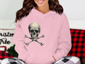 A person in a pink Skull and Crossbones hoodie by Garment Graphics sits with plaid pants, while a Christmas-themed pillow adds festive comfort to the background.